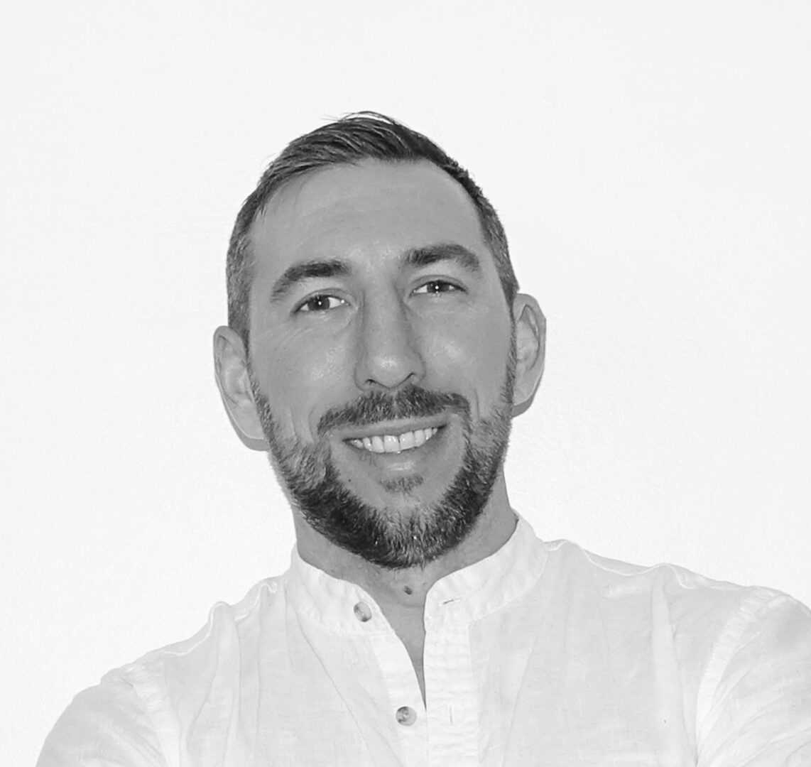 Uomo sorridente con barba e capelli corti, indossa una camicia bianca, sfondo neutro.